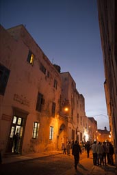 Essaouira, small street light lit alley at night, people strolling, sky dark blue.