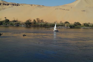 Lone felucca, blue Nile at Aswan, desert dunes in back.
