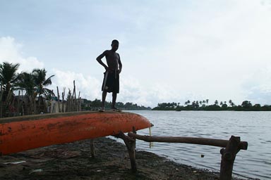 Harper, Liberia, African child on top of pirogue, harbor in back, black against sun.
