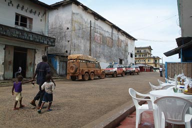 Harper, Maryland, Liberia, sophie's spot cafe in mechlin street, 6x6 Land Rover Defender parked, African father and children walk by, white plastic chairs and tables, 2 white and red UN Police Land Cruisers parked next to Land Rover.